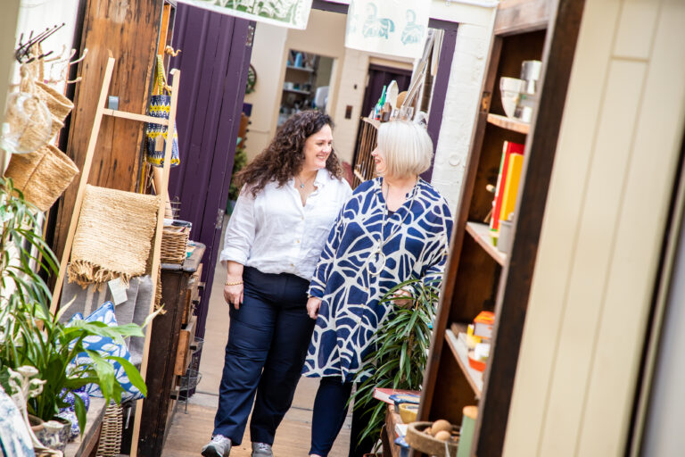Two women smiling at each other walking in corridor with bookshelves and plants